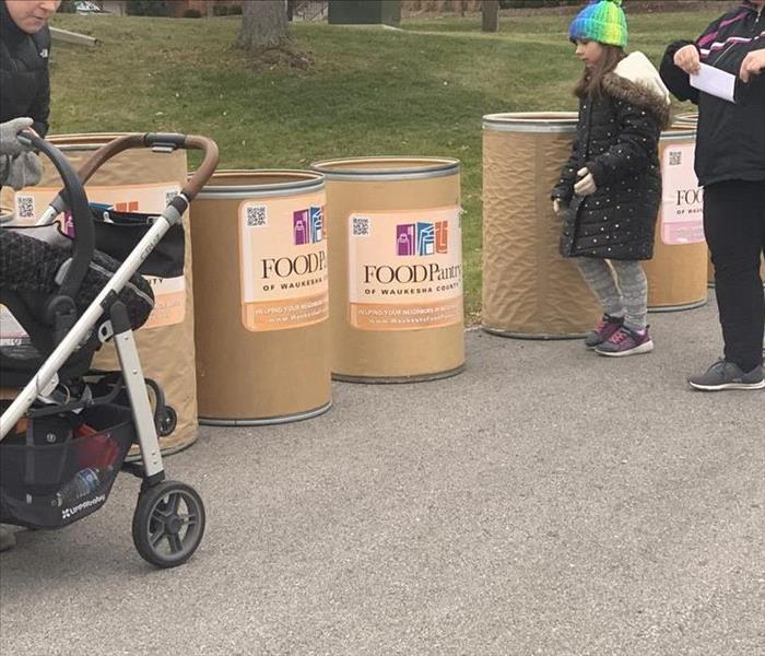 1 child and 2 adults donating food into round food bins with signs that say Waukesha County Food Pantry 